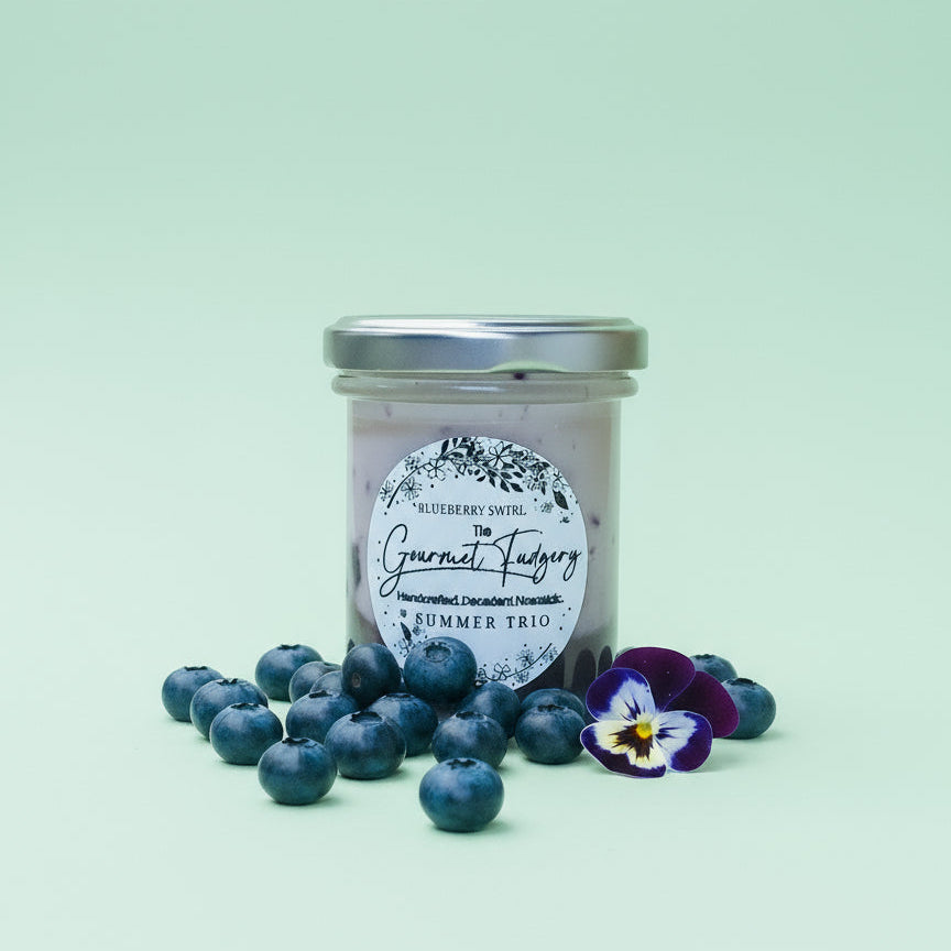 Jar of 'Blueberry Swirl' Gourmet Fudge from 'The Gourmet Fudgery' with a floral label on a mint coloured background and blueberries in the foreground.